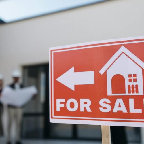 A real estate sign indicates a property for sale as two agents in hard hats discuss building plans outdoors.